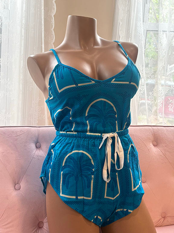 Mannequin wearing a blue Kilo Brava cotton cami and short set with a white palm and arch print, displayed on a blush pink tufted bench near a window. The set features adjustable spaghetti straps, a drawstring waistband, and curved hems.